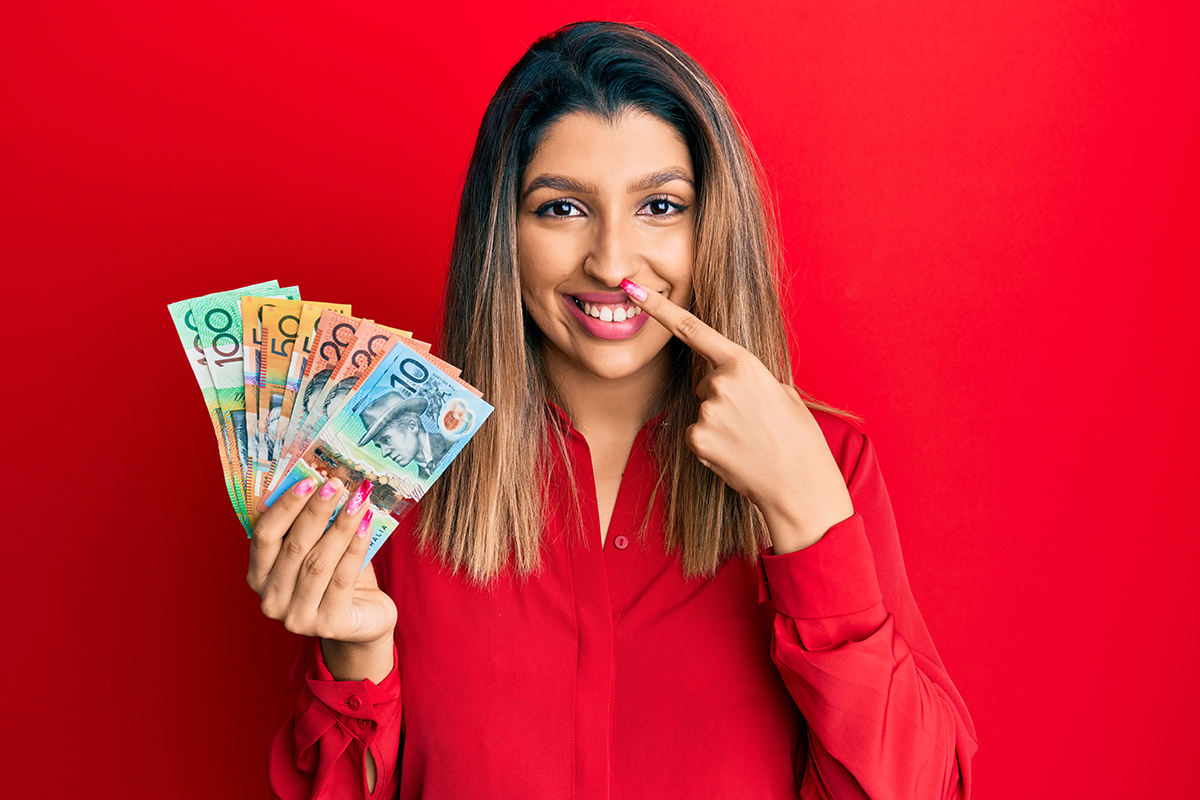 beautiful brunette woman holding dollars pointing with hand finger to face and nose smiling cheerful
