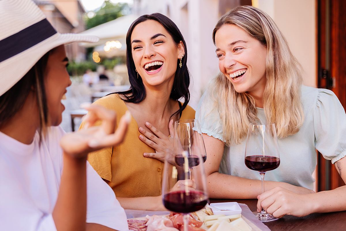 three happy diverse women having fun enjoying aperitif with red wine sitting at an outside table in a bar