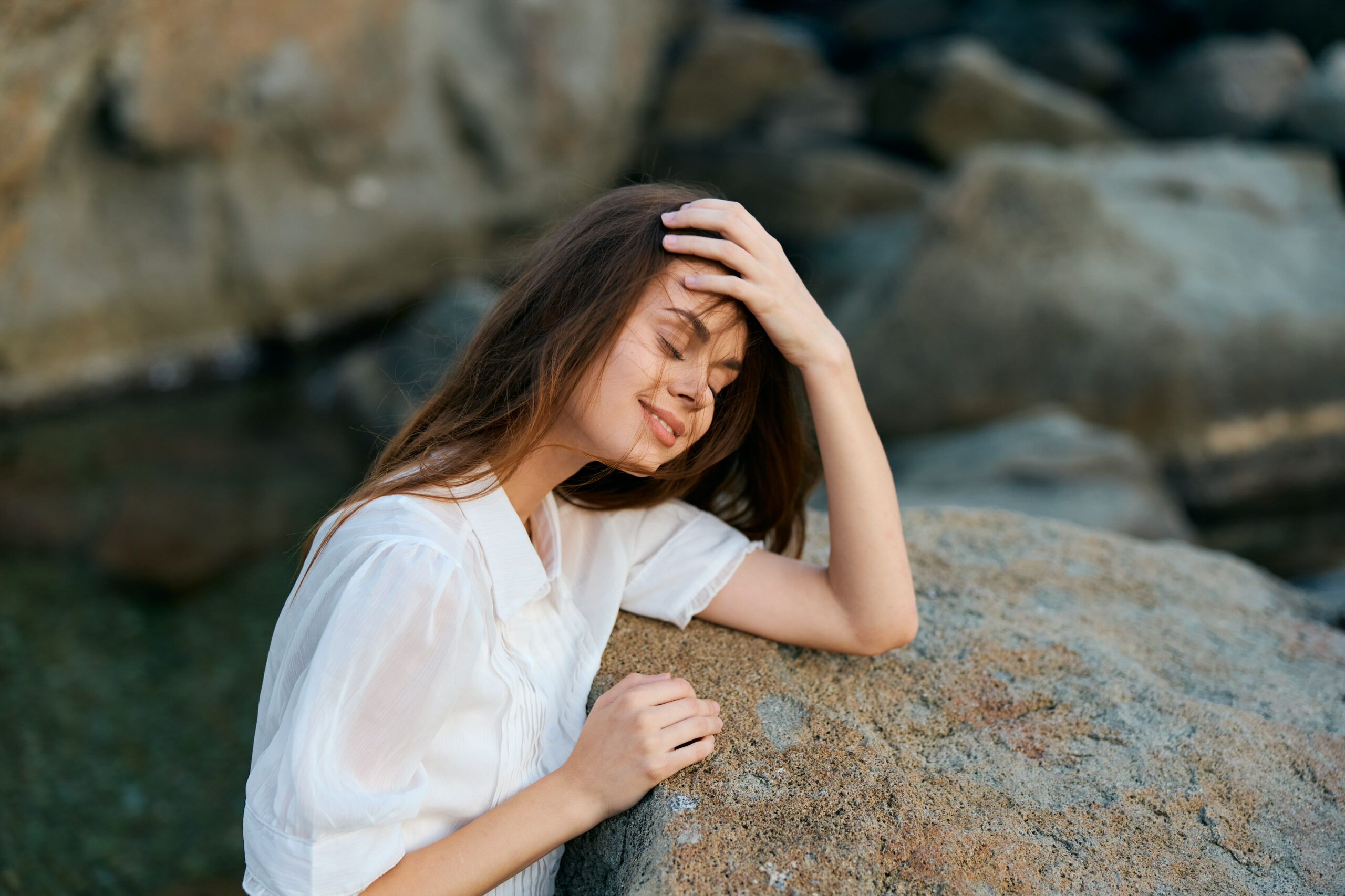 Serene woman sitting on ocean rock with hands on head, gazing at camera in peaceful contemplation