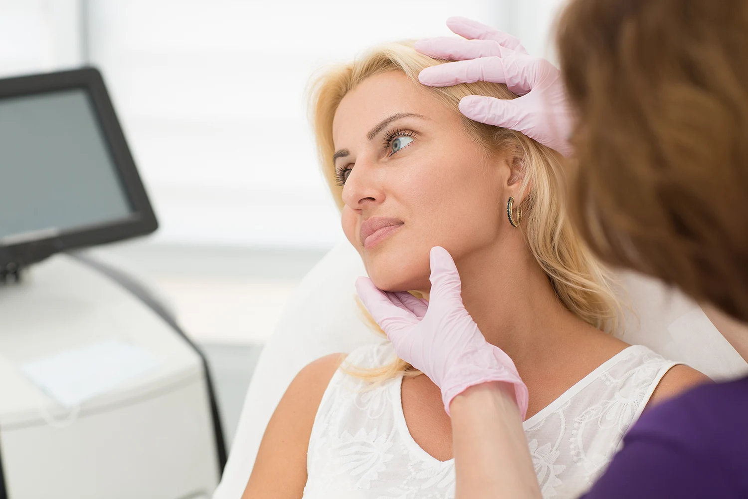 Aesthetic professional in pink gloves examining a blonde woman’s facial skin in a modern medical clinic. Aesthetic professional in pink gloves examining a blonde woman's facial skin in a modern medical clinic.