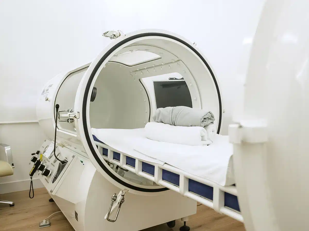 Modern monoplace hyperbaric oxygen therapy chamber with white bedding and control panel in a medical clinic.